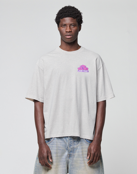 A young man with short curly hair wears the LIVE FAST DIE YOUNG LFDY Real Ones Tee, an oversized light gray 100% Baumwolle T-shirt with pink and purple chest text, and light blue jeans, standing against a plain white background.