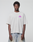A young man with short curly hair wears the LIVE FAST DIE YOUNG LFDY Real Ones Tee, an oversized light gray 100% Baumwolle T-shirt with pink and purple chest text, and light blue jeans, standing against a plain white background.