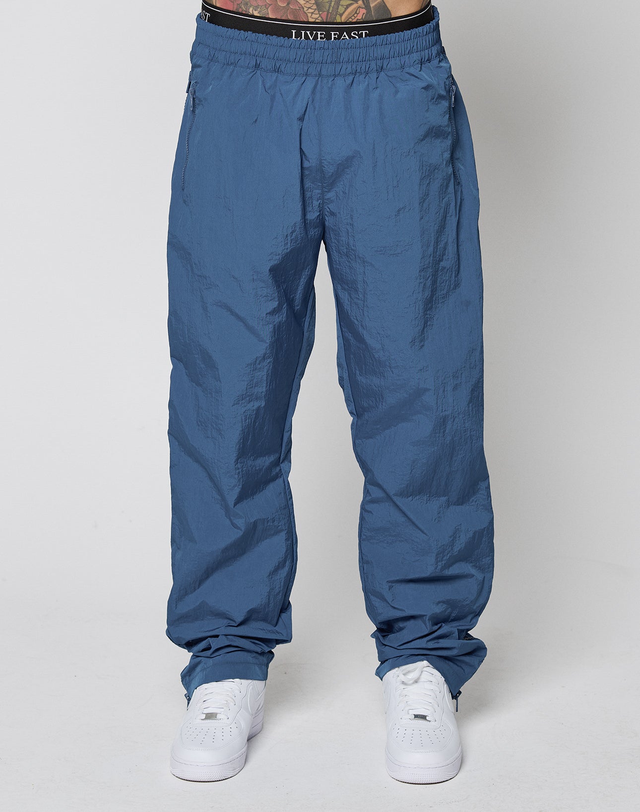 A person wearing loose blue Zip Tape Track Pants by LIVE FAST DIE YOUNG with an elastic waistband and white sneakers stands against a plain background. The waistband says LIVE FAST, showing a hint of tattooed skin above the pants.