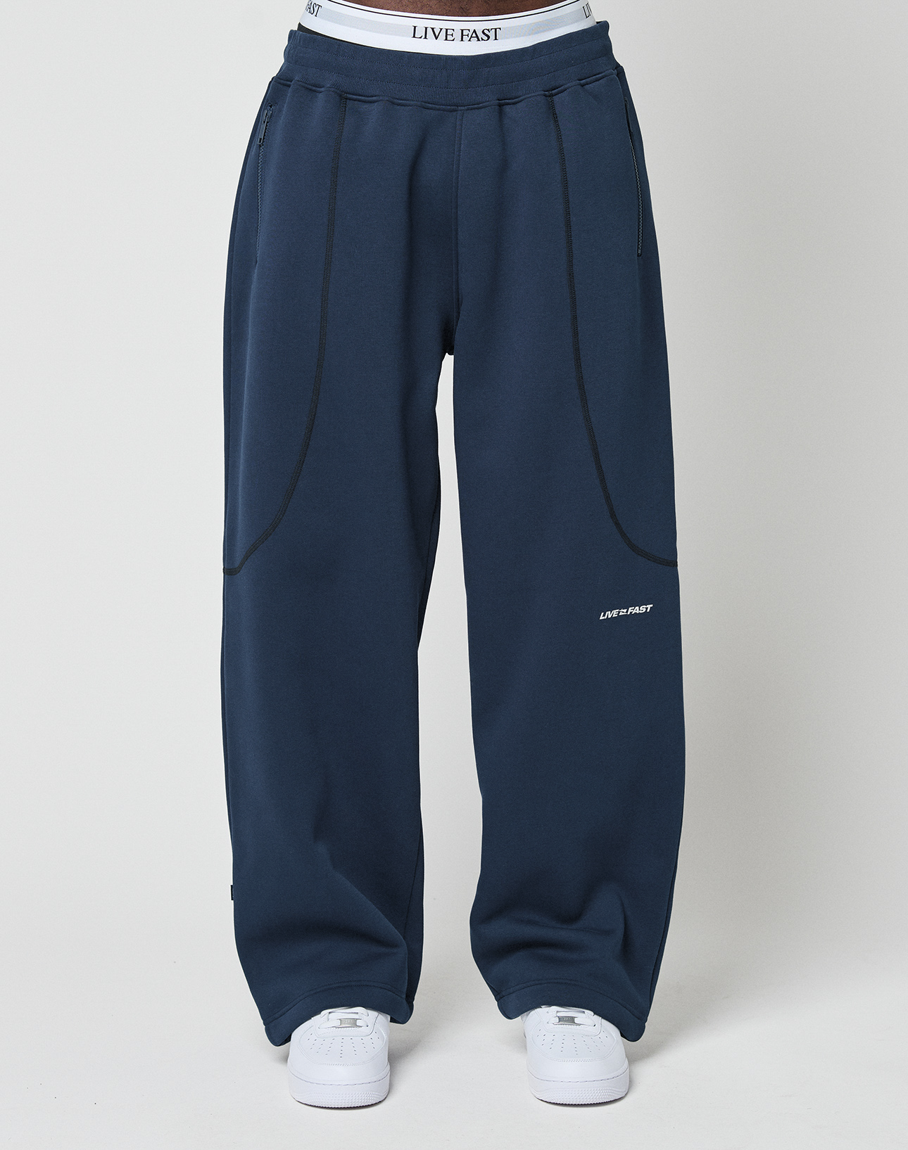 Someone wearing LFDY Active Fleece Jogger by LIVE FAST DIE YOUNG in navy blue with black seam details, a visible LIVE FAST logo waistband, and white sneakers, standing against a plain light background.