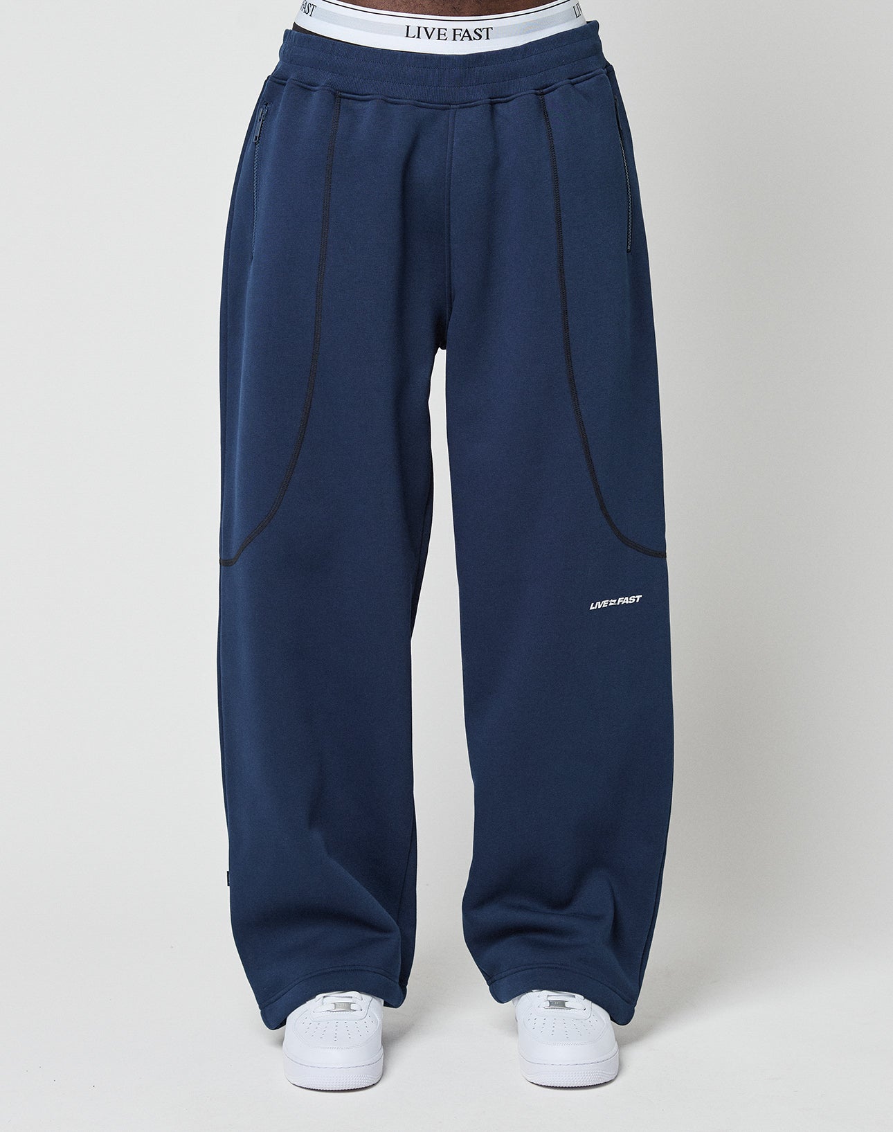Person wears loose-fitting navy blue LFDY Active Fleece Jogger by LIVE FAST DIE YOUNG, with black seam details, white LIVE FAST logo on left thigh, zip pockets, white sneakers, and visible waistband of white LIVE FAST underwear.