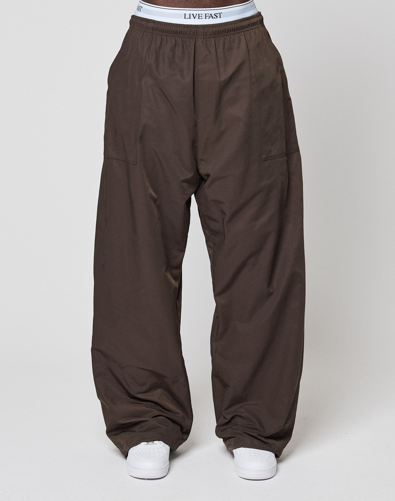 A person wears wide fit Baggy Nylon Pants by LIVE FAST DIE YOUNG and white athletic shoes, with the LIVE FAST-branded white underwear's waistband visible above the pants. The background is plain and light-colored.