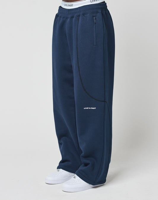 Person wearing LFDY Active Fleece Jogger by LIVE FAST DIE YOUNG—wide fit navy sweatpants made from soft Baumwolle with side zip pocket, curved seams, LIVE FAST logo on thigh, white waistband, and white sneakers—standing against a light background. | model