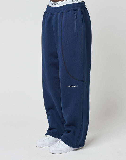 A person wears the LFDY Active Fleece Jogger by LIVE FAST DIE YOUNG in wide fit navy with black piping, LIVE FAST logo on waistband and thigh, Reißverschluss Taschen, and white sneakers. The background is plain white.