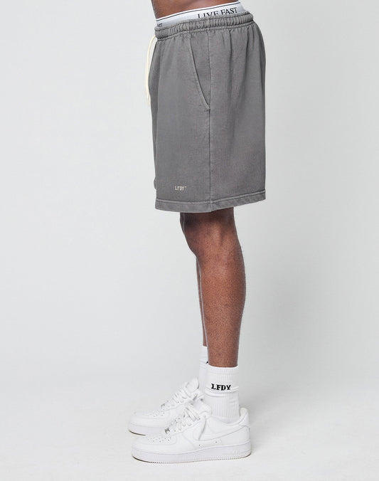 A person in profile wears LIVE FAST DIE YOUNG Basic Sweat Shorts with elastic waistband, featuring LIVE FAST text, paired with white LFDY socks and sneakers. The regular fit shorts are gray against a plain light background.