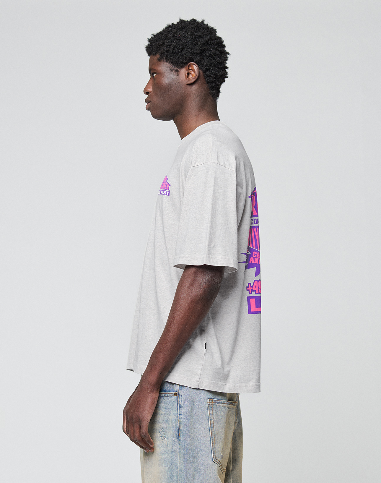 A young man in profile wears the LFDY Real Ones Tee by LIVE FAST DIE YOUNG—an oversize light gray 100% cotton T-shirt with purple back graphics and a Flaglabel detail—paired with light blue jeans against a white background.