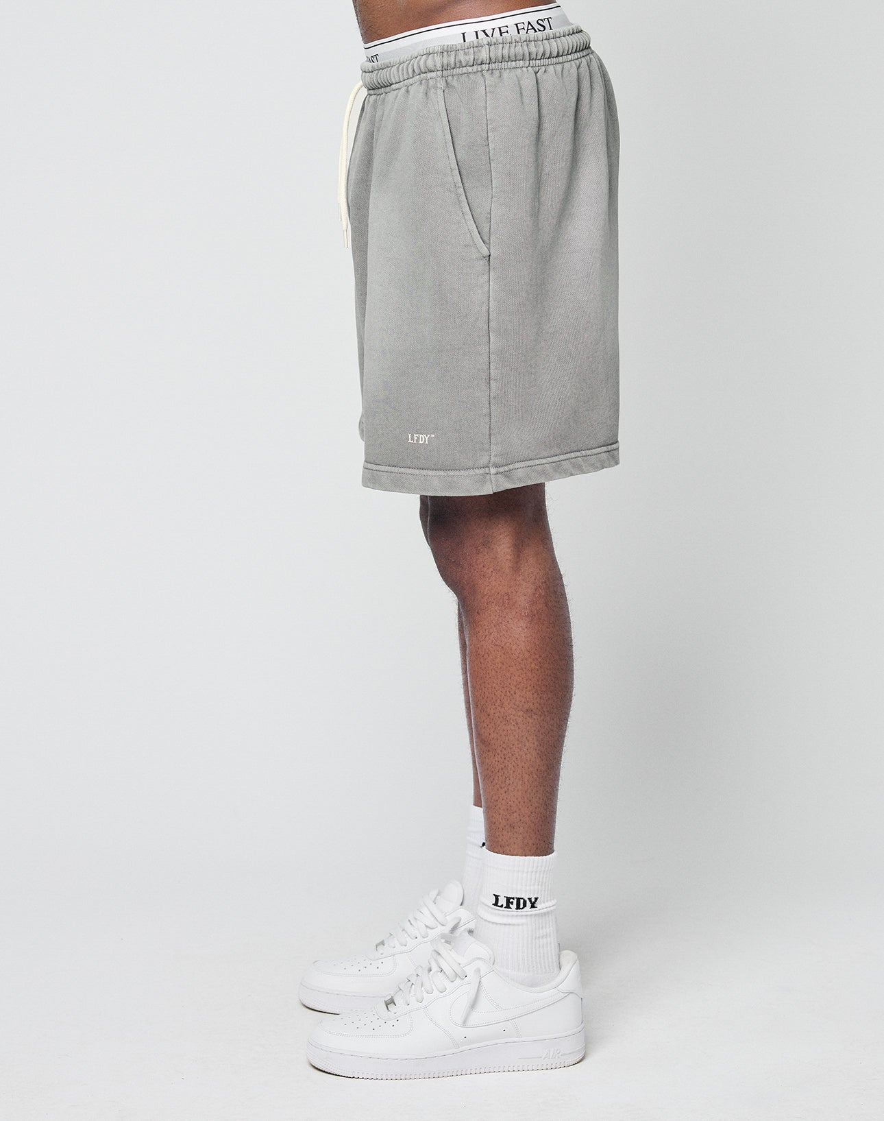 Person standing sideways in LIVE FAST DIE YOUNG Basic Sweat Shorts (gray, faded), with waistband showing LIVE FAST text and a small LFDY logo near the hem. Worn with white LFDY socks and white Nike sneakers. Plain white background.