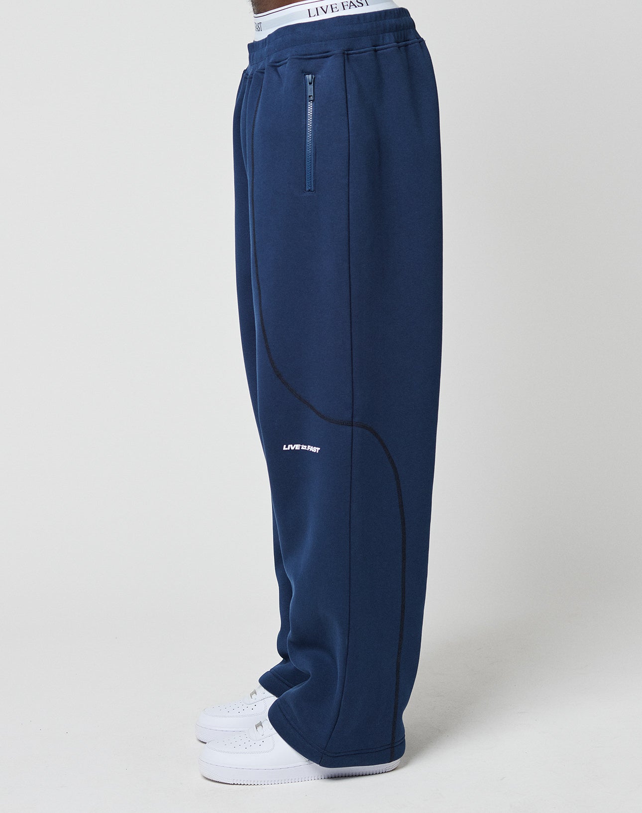 A person in loose, dark blue LFDY Active Fleece Jogger by LIVE FAST DIE YOUNG with Reißverschluss Taschen, curved seam, bold LIVE Logo Print on the left leg, and white sneakers, shown from the side.