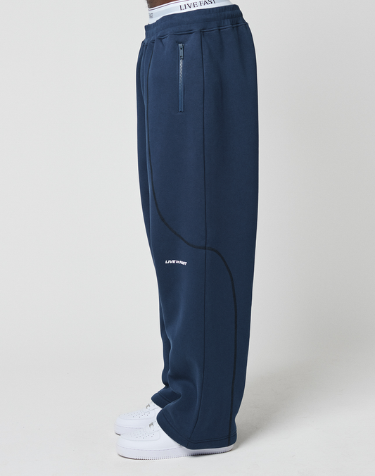 A person in wide fit navy LFDY Active Fleece Jogger by LIVE FAST DIE YOUNG, featuring black seam details, zippered pocket, and a small LIVE FAST logo near the knee, styled with white sneakers and posed sideways against a white background.