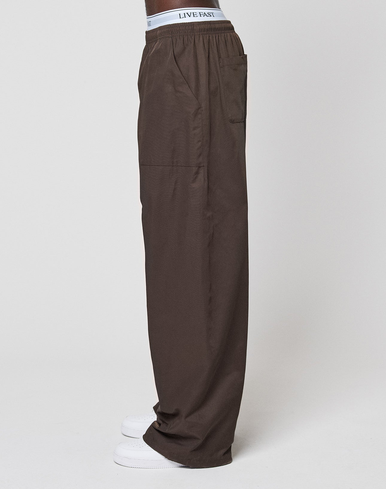 A person wearing LIVE FAST DIE YOUNG Baggy Nylon Pants with an elastic waistband, visible LIVE FAST branding, wide-leg fit, paired with white sneakers, standing in profile against a plain background for a relaxed everyday look.