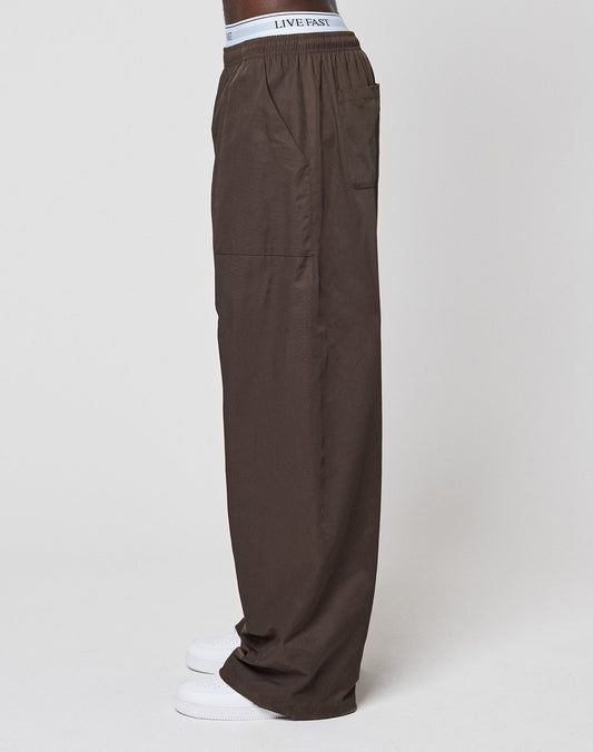 A person wearing LIVE FAST DIE YOUNG Baggy Nylon Pants with an elastic waistband, visible LIVE FAST branding, wide-leg fit, paired with white sneakers, standing in profile against a plain background for a relaxed everyday look.
