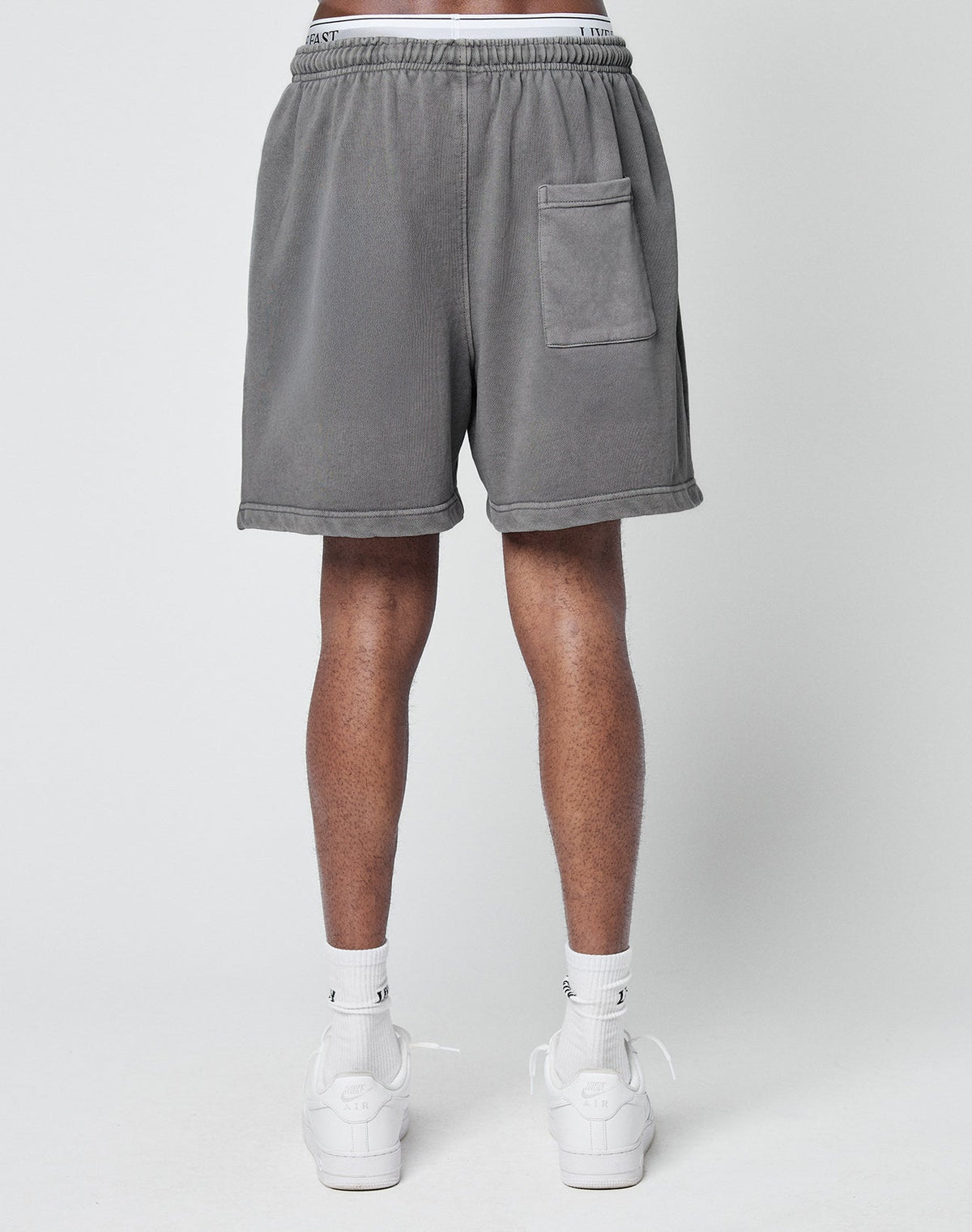 A person shown from the back against a plain light background wears LIVE FAST DIE YOUNG Basic Sweat Shorts in gray with an elastic waist and side pocket, paired with white socks and white sneakers.