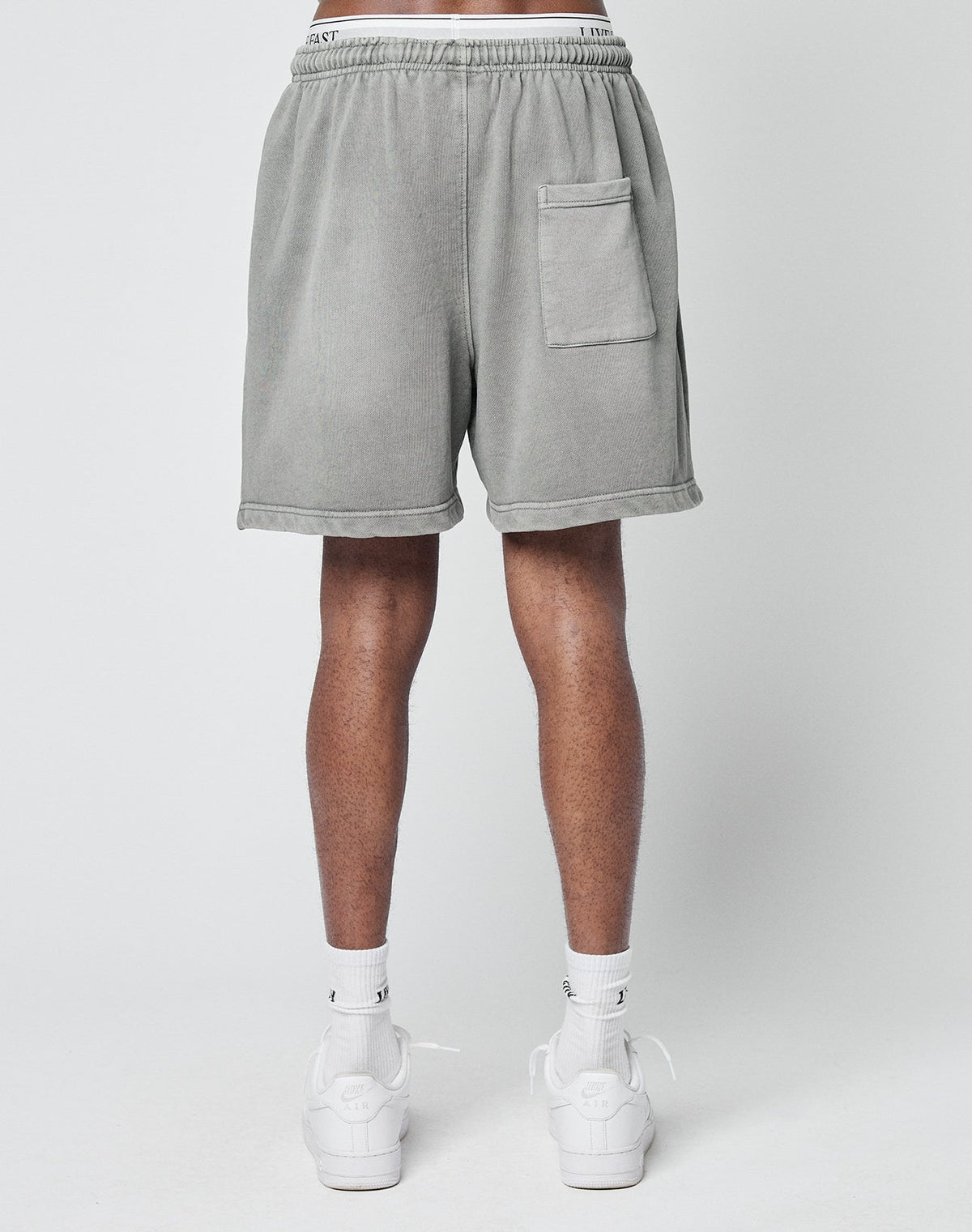 A person models LIVE FAST DIE YOUNG Basic Sweat Shorts in light gray, a regular fit with a back pocket, paired with white socks and sneakers, standing against a plain light gray background, shown from the back waist down.