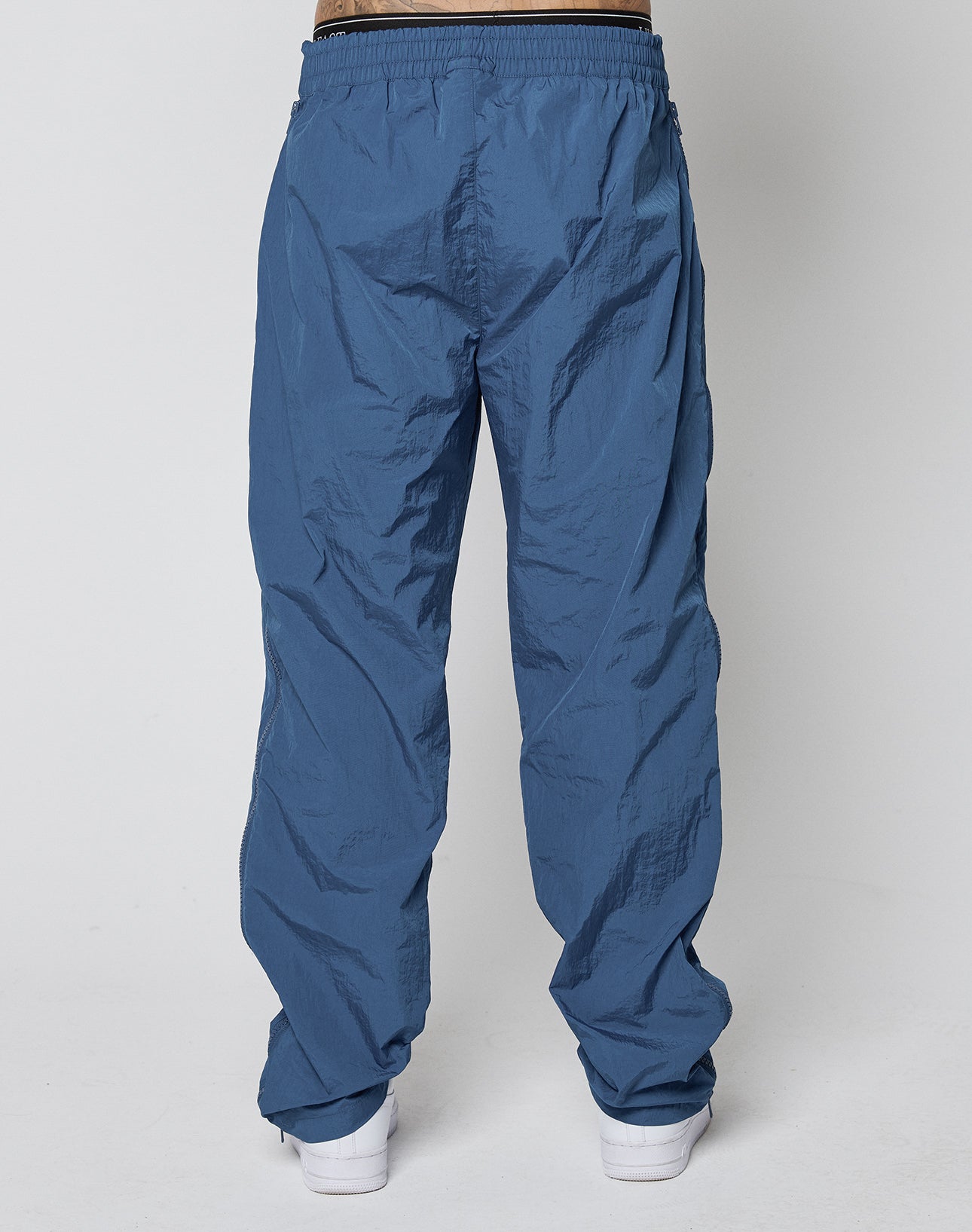 Someone wearing LIVE FAST DIE YOUNG Zip Tape Track Pants with an elastic waistband and white sneakers, shown from the back against a plain light gray background.