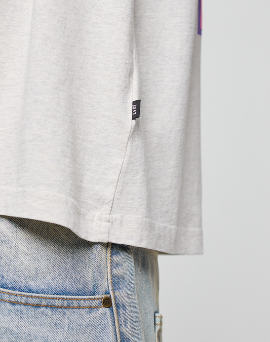 Close-up of the LIVE FAST DIE YOUNG LFDY Real Ones Tee in light gray, 100% cotton, featuring a small black flag label and oversize fit, worn over light blue jeans with only the side of the shirt and jeans pocket visible.