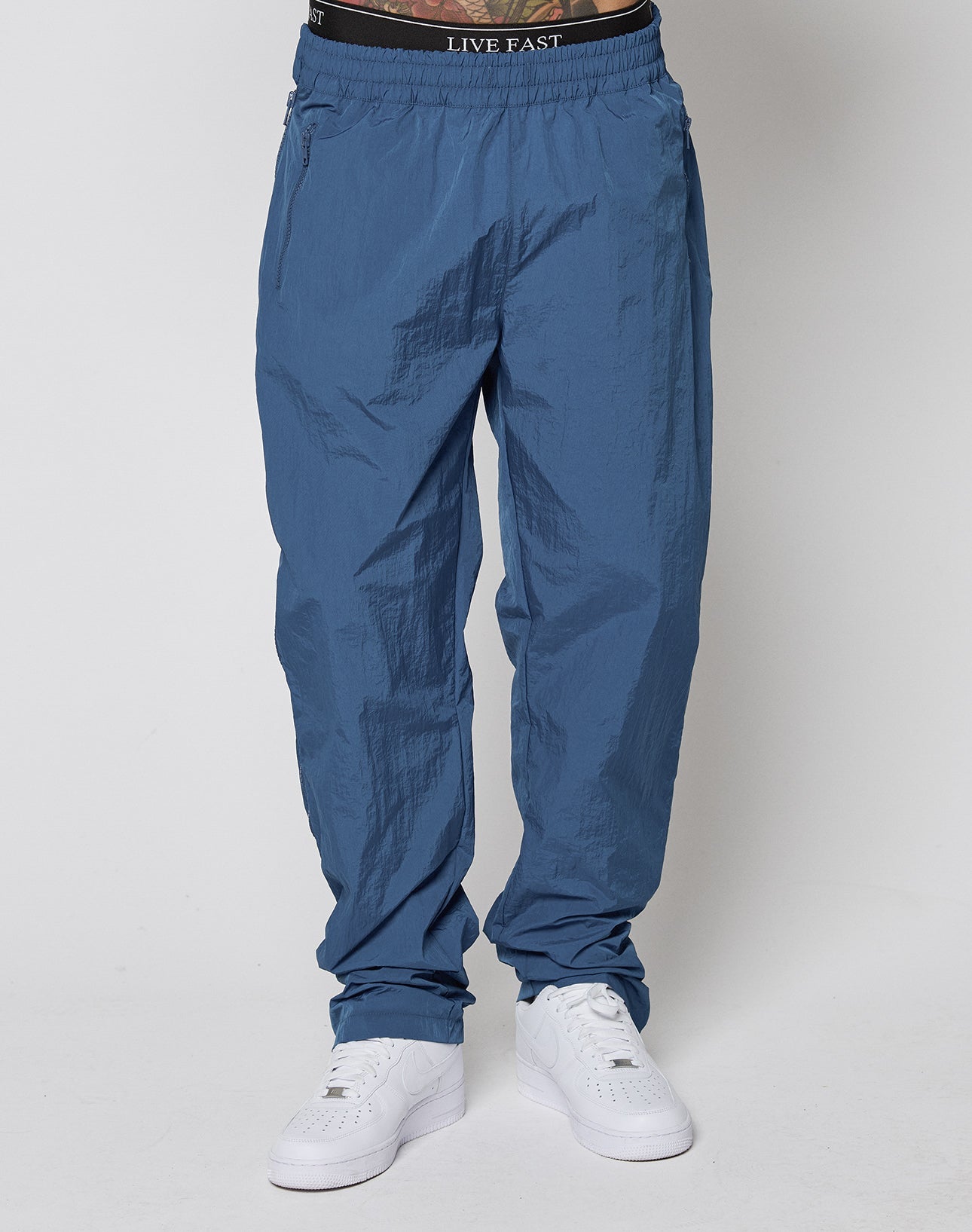 A person wears blue LIVE FAST DIE YOUNG Zip Tape Track Pants with an elastic waistband, white sneakers, and a visible black LIVE FAST waistband, standing against a plain white background. Only the lower body is shown.