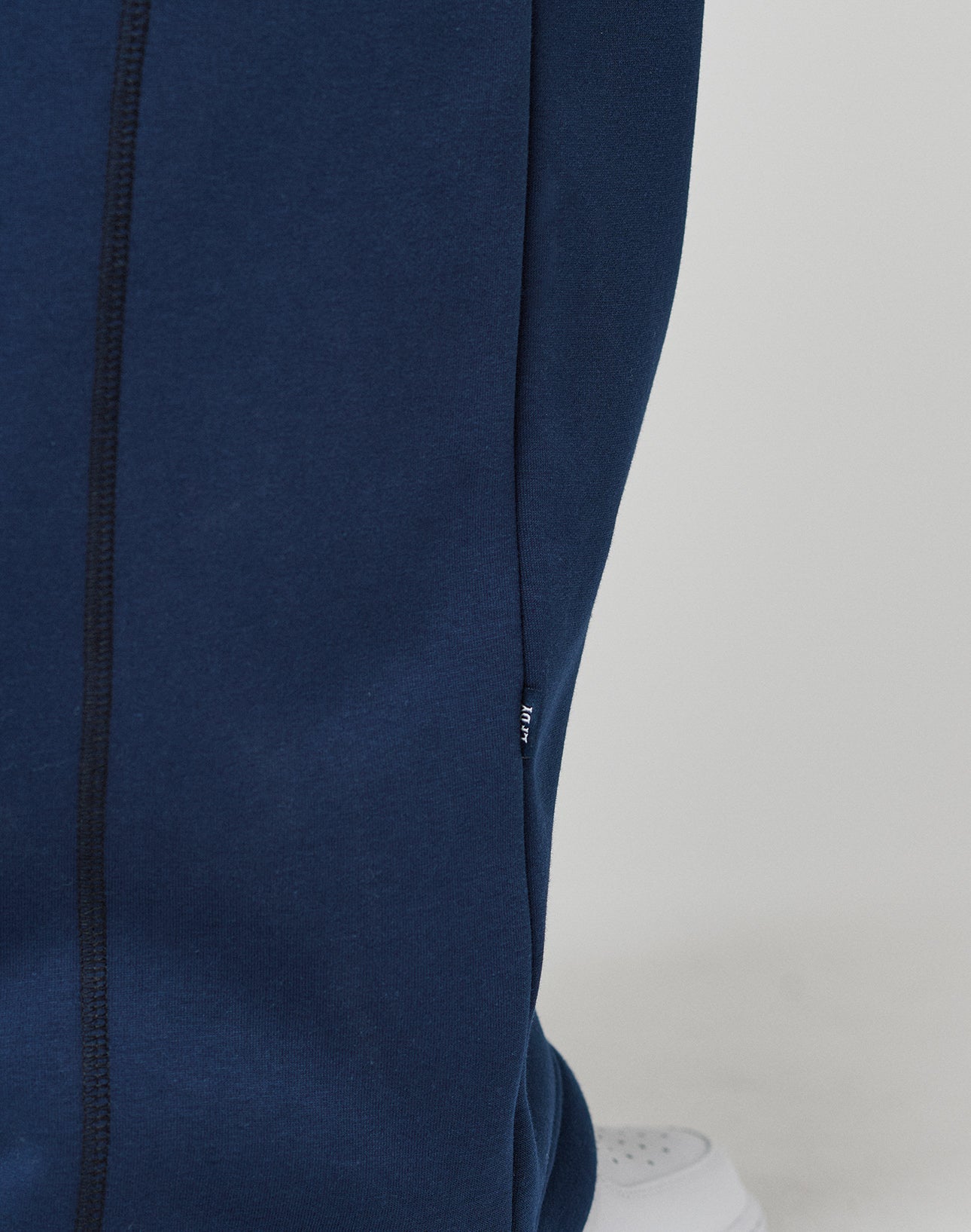 Close-up of a person wearing navy blue LFDY Active Fleece Jogger by LIVE FAST DIE YOUNG, featuring a visible seam, small black tag, and logo print. Part of a white sneaker is seen at the bottom against a plain light background.
