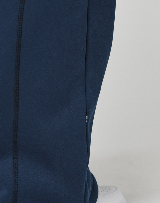 Close-up of a person wearing LIVE FAST DIE YOUNG's LFDY Active Fleece Jogger, dark blue wide fit pants with a visible seam and small fabric tag. A white sneaker is partially visible on a light surface.
