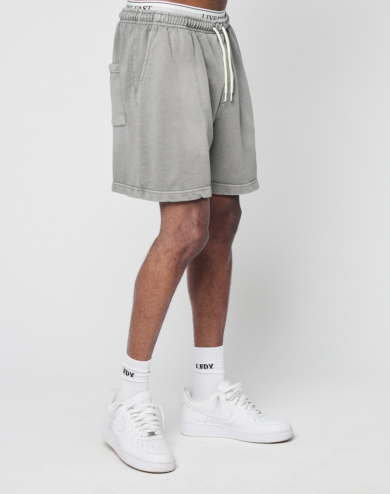 Only the lower half of the body is visible as a person stands against a white background, wearing LIVE FAST DIE YOUNG Basic Sweat Shorts in light gray, paired with white crew socks featuring an LFDY logo and white sneakers.