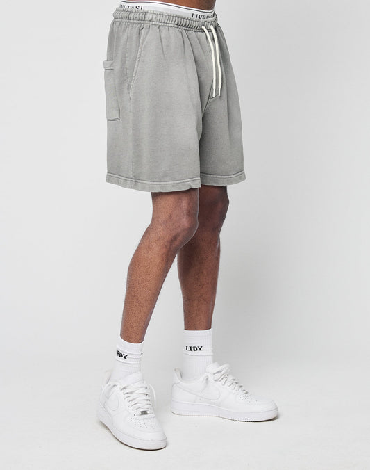 Only the lower half of the body is visible as a person stands against a white background, wearing LIVE FAST DIE YOUNG Basic Sweat Shorts in light gray, paired with white crew socks featuring an LFDY logo and white sneakers.