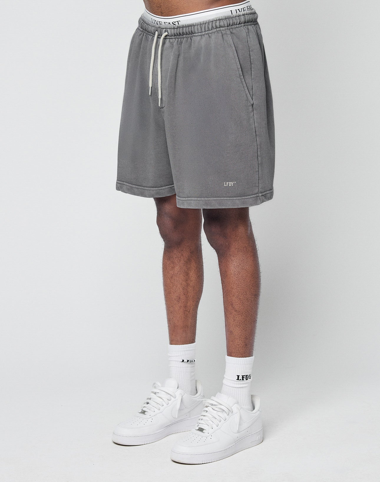 A person wears LIVE FAST DIE YOUNG Basic Sweat Shorts in gray with elastic waistband, side pockets, drawstring, and logo print. The regular fit and LFDY-branded waistband ensure comfort, all shown against a plain white background. | model