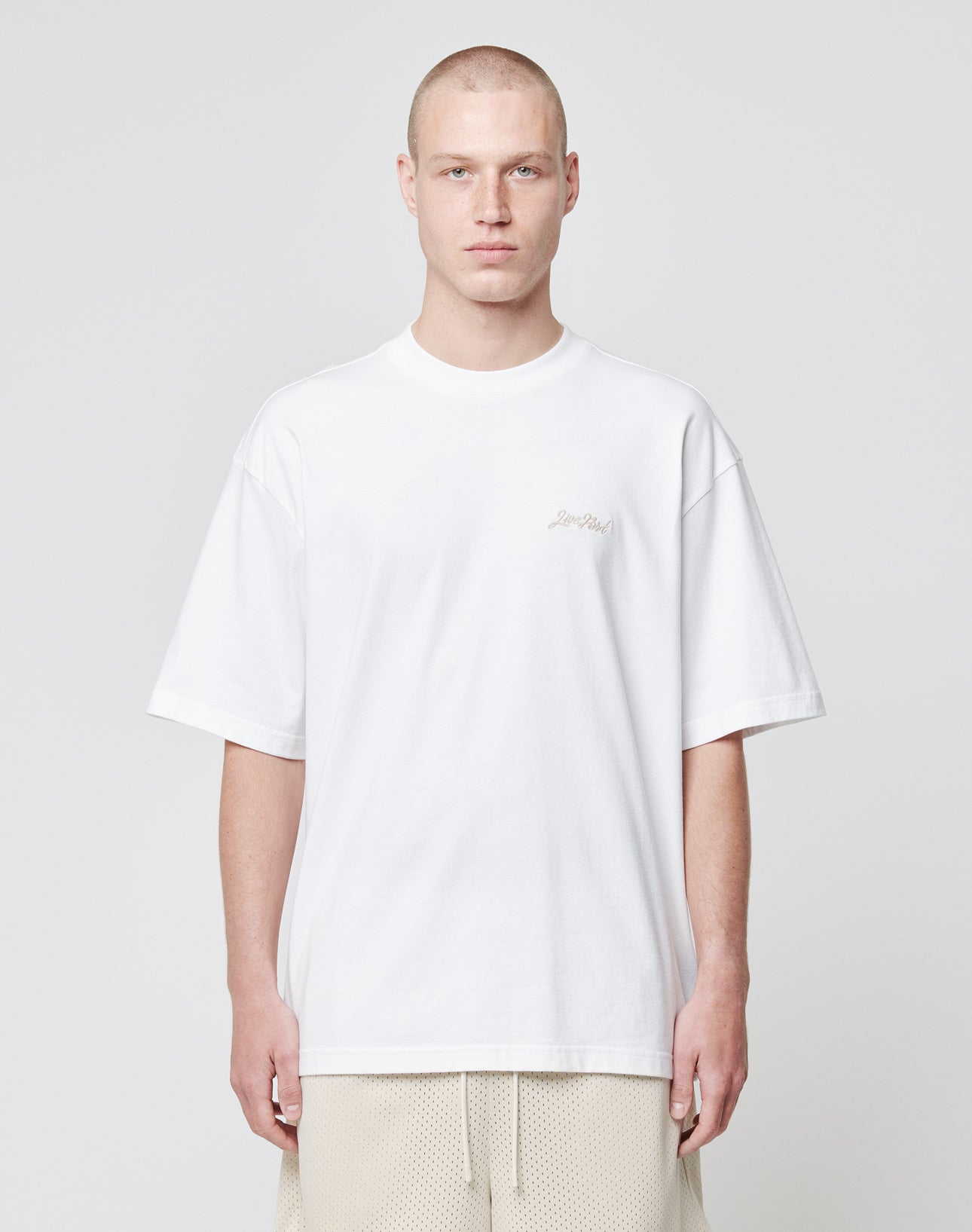 A person with a shaved head wears the LIVE FAST DIE YOUNG Basic Script Tee in plain white and beige shorts, standing against a light grey background, looking directly at the camera with a neutral expression.