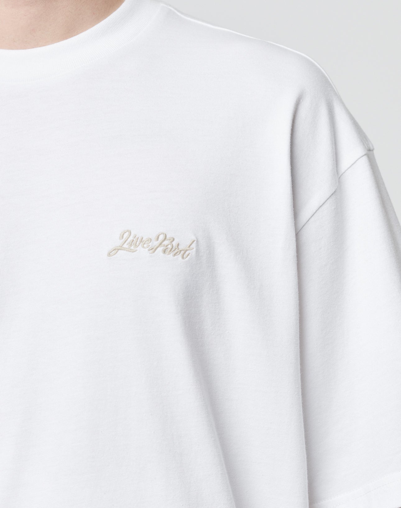 Close-up of a person wearing the LIVE FAST DIE YOUNG Basic Script Tee, an oversize plain white T-shirt made from 100% Baumwolle, with Live Past embroidered in small, light-colored text on the upper left chest.