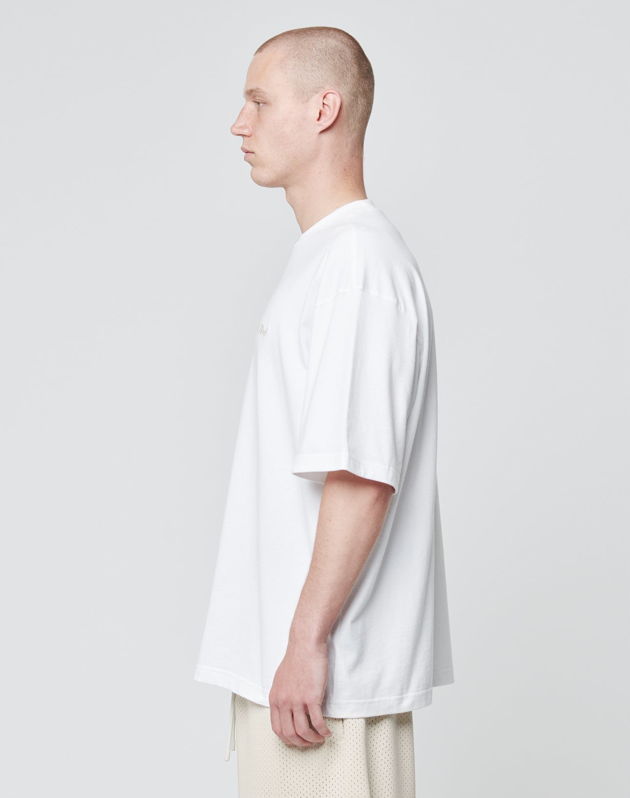 A man with a shaved head stands in profile against a plain background, wearing the LIVE FAST DIE YOUNG Basic Script Tee, an oversize fit white T-shirt made from 100% Baumwolle, paired with light-colored shorts.