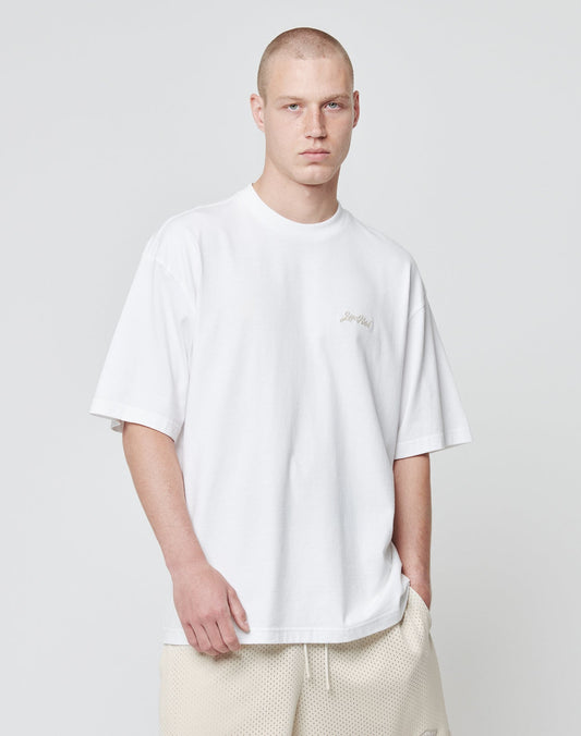 A young man with a shaved head wears the LIVE FAST DIE YOUNG Basic Script Tee in an oversize fit and beige shorts, standing against a plain light gray background, looking directly at the camera with a neutral expression.