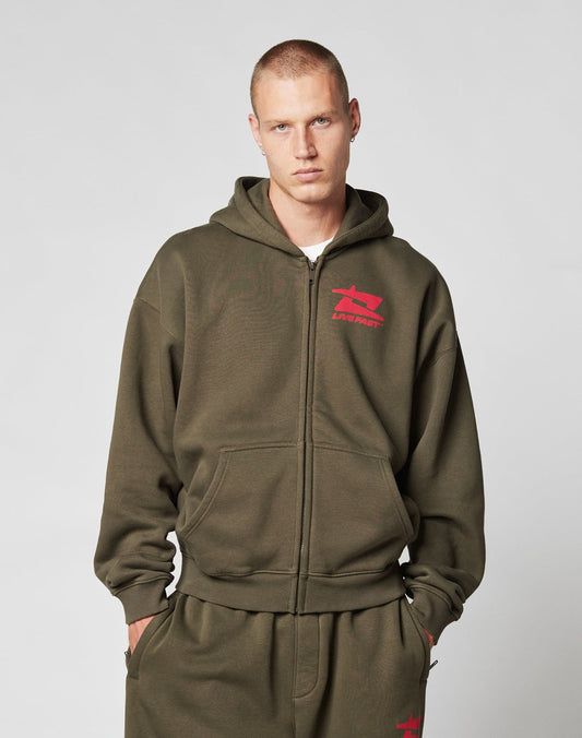 A young man with short hair wears the LIVE FAST DIE YOUNG Live Fast Sweatsuit Jacket in olive green, styled oversized with bold red front-print logos, and matching sweatpants, standing hands-in-pockets against a plain light background.