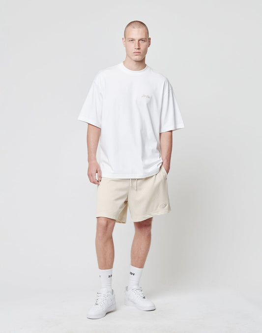 A man with short hair and a neutral expression stands against a white background wearing LIVE FAST DIE YOUNG Basic Mesh Shorts in light beige, a loose Regular Fit white T-shirt, white socks, and white sneakers, with his left hand in his pocket.