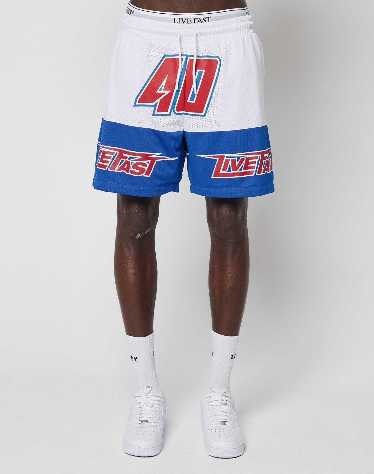 A person models LFDY Racing Mesh Shorts by LIVE FAST DIE YOUNG—regular fit white polyester shorts with a bold red 40, blue LIVE FAST stripes, paired with white socks and sneakers against a plain white background.