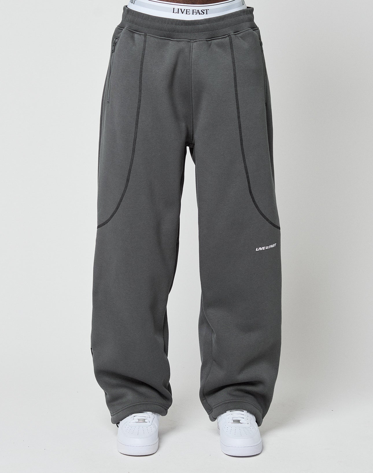 A person wears LFDY Active Fleece Jogger by LIVE FAST DIE YOUNG in wide fit, dark gray with subtle seams and flatlock-topstitching, paired with white sneakers. The waistband of LIVE FAST logo underwear is visible above the joggers.