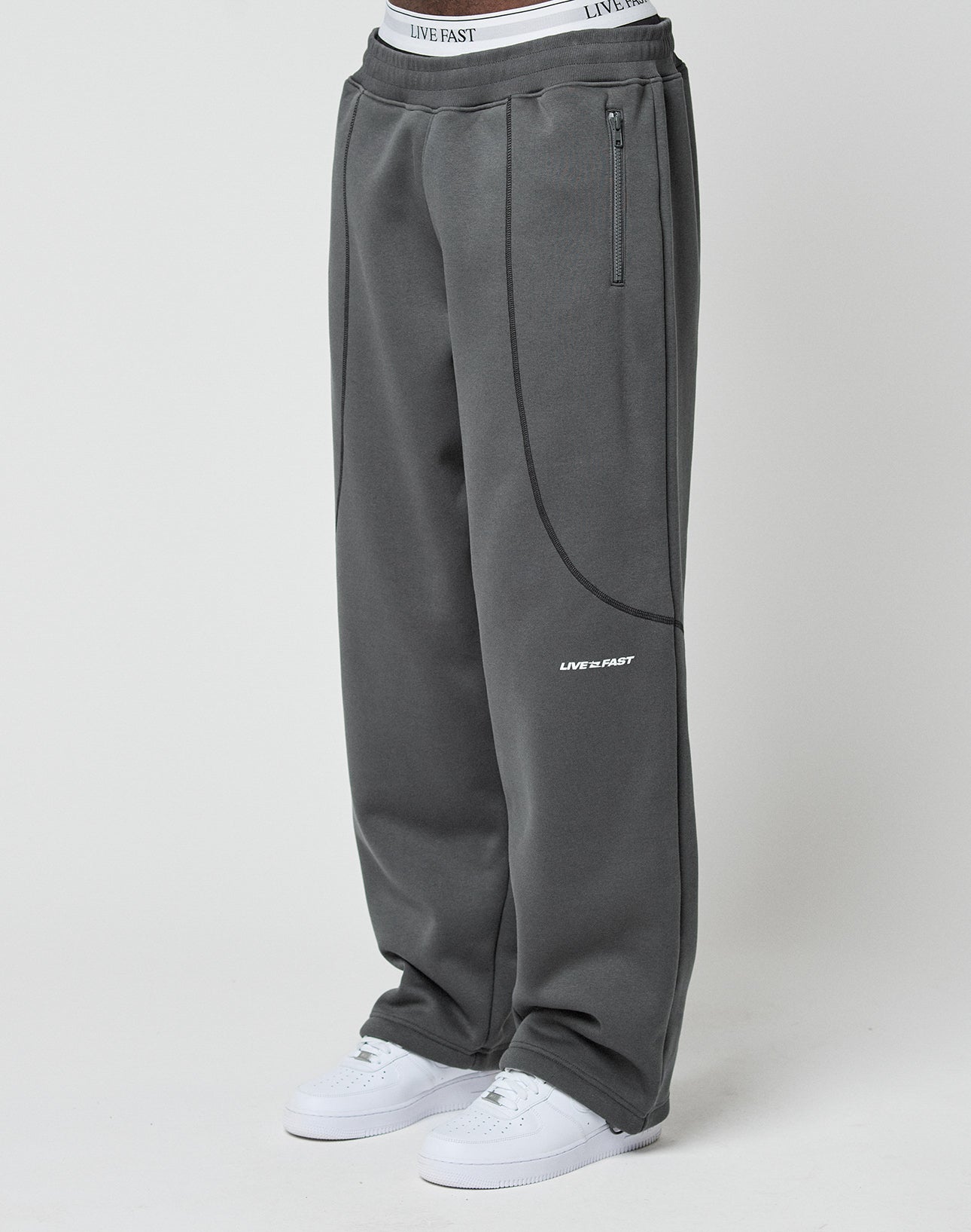 A person wears the LFDY Active Fleece Jogger by LIVE FAST DIE YOUNG in dark gray with zip pockets, a waistband featuring LIVE FAST text, and a small LIVE FAST logo on one leg, paired with white sneakers. | model