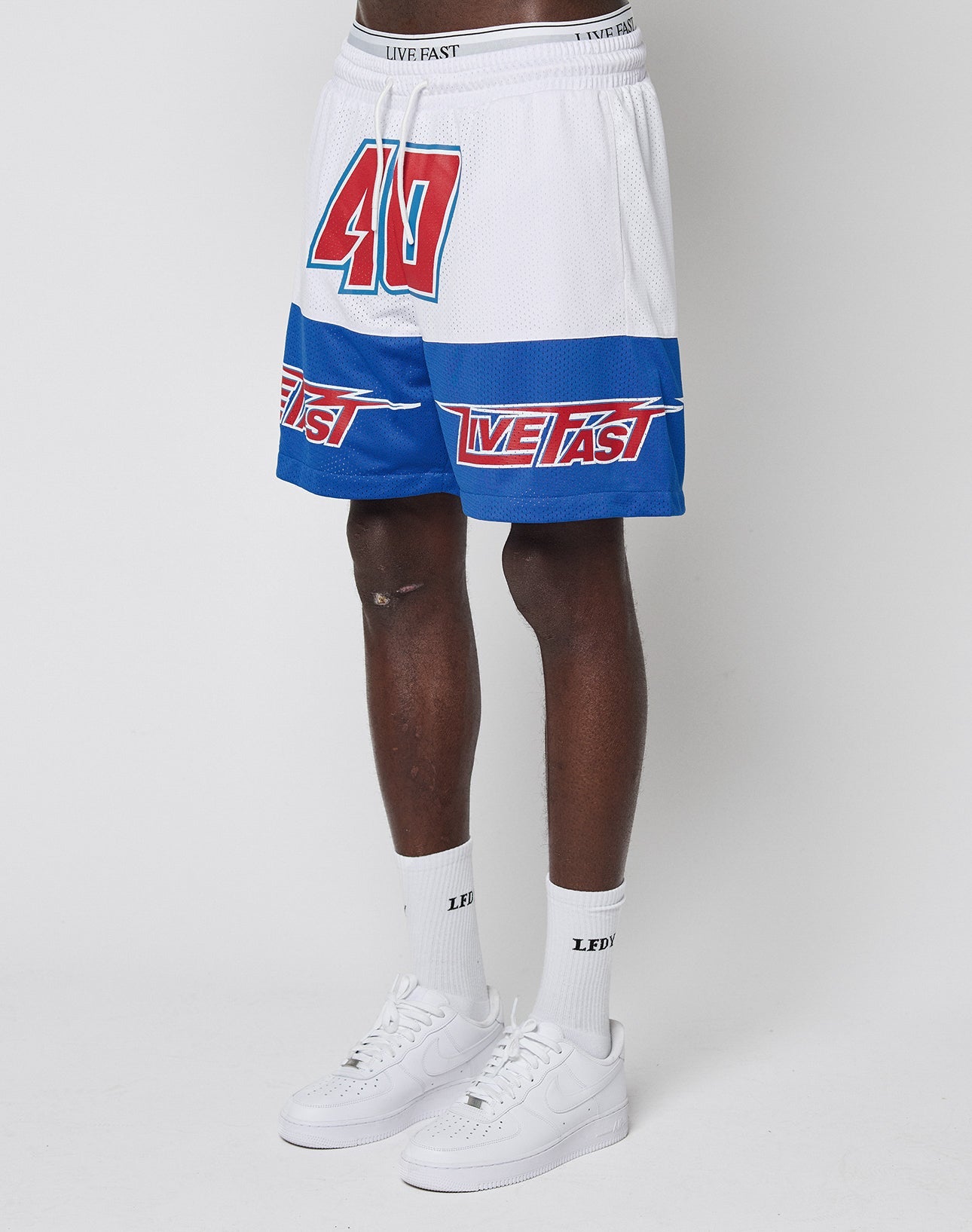 A person wears LFDY Racing Mesh Shorts by LIVE FAST DIE YOUNG, featuring bold prints, a red 40, and LIVE FAST text, paired with white LFDY socks and Nike Air Force 1s, standing against a plain white background.
