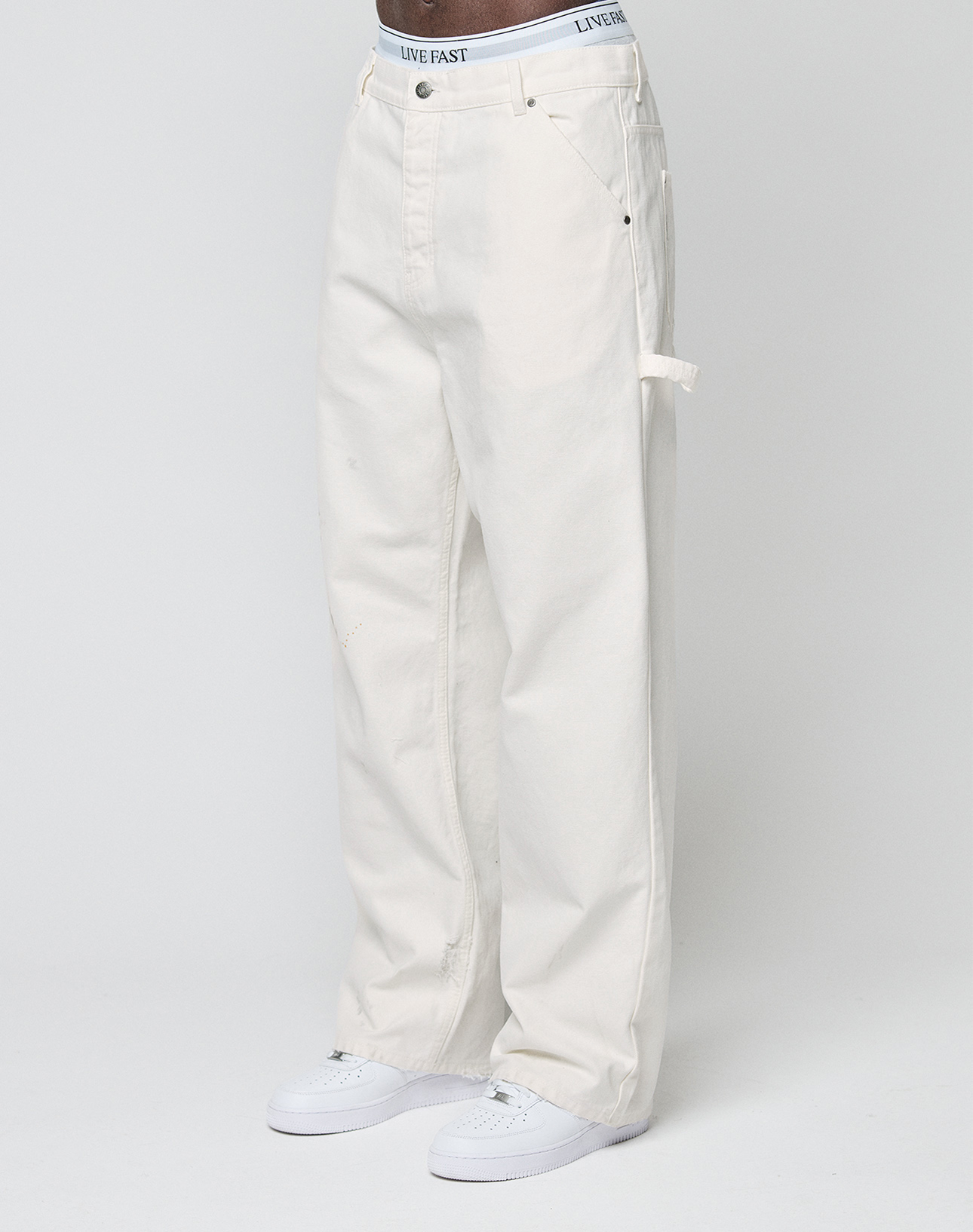 A person models LIVE FAST DIE YOUNG Worker Pants—loose-fit, off-white carpenter style with a carpenter pocket—paired with white sneakers and a waistband featuring LIVE FAST, standing against a plain light background. | model