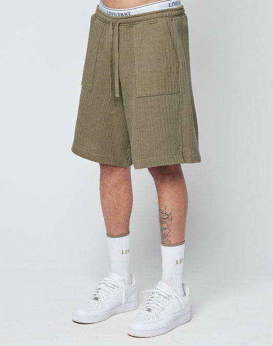 A person wears LIVE FAST DIE YOUNG Basic Knit Shorts in olive green with an elastic waistband, white LIVE FAST waistband underwear, white socks featuring LIVE FAST text, and white Nike sneakers against a plain light background.