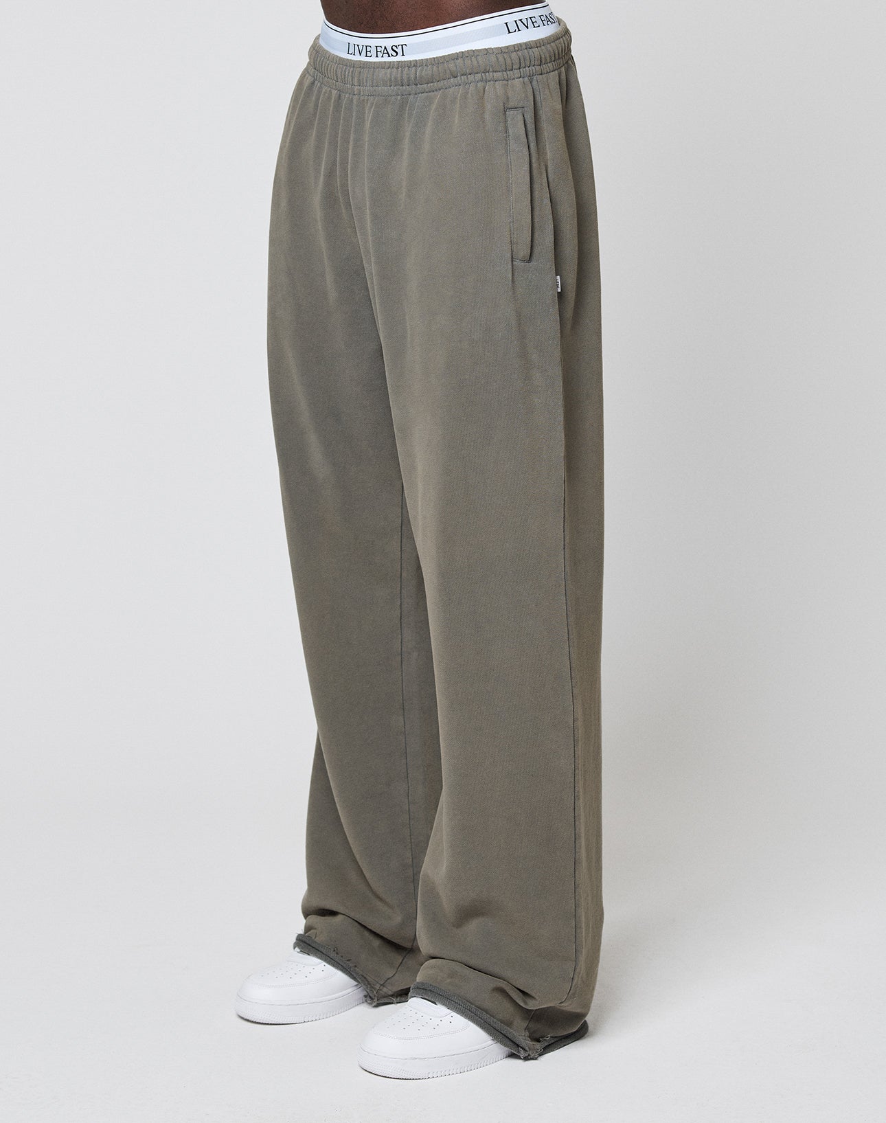A person wears LFDY Ripped Sweatpants by LIVE FAST DIE YOUNG in olive green with a wide fit and elastic waistband, paired with white sneakers and LIVE FAST waistband underwear, standing against a plain light background.