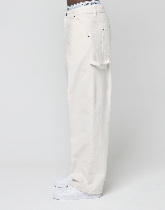 A person stands sideways in off-white LIVE FAST DIE YOUNG Worker Pants, 100% cotton, loose fit with a visible branded waistband and carpenter pocket, paired with white sneakers against a plain background.