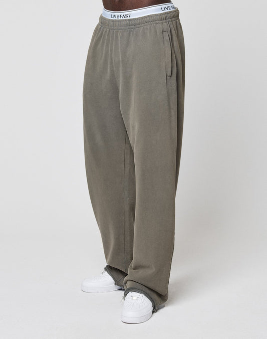 A person is standing against a plain white background, wearing LFDY Ripped Sweatpants from LIVE FAST DIE YOUNG in olive green wide fit with an elastic waistband, paired with white sneakers.