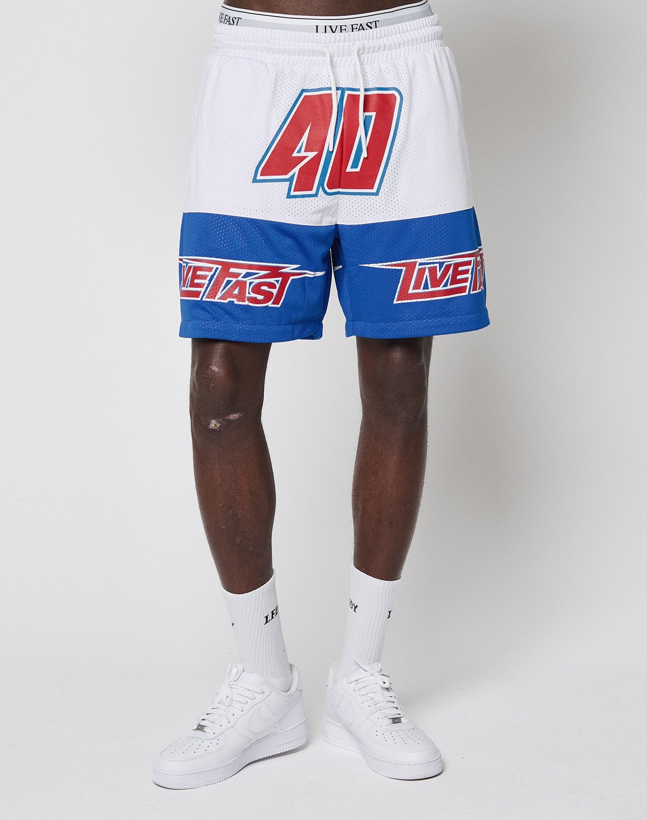 A person wears LFDY Racing Mesh Shorts by LIVE FAST DIE YOUNG, white and blue with a large red 40 on the left leg and LIVE FAST across the front, featuring bold prints, paired with white sneakers and socks in a regular fit.