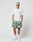 A man stands against a plain background wearing the LIVE FAST DIE YOUNG LFDY Thunder Chest Tee in white, paired with green and white tie-dye shorts, a matching cap, white crew socks, and sneakers. He looks straight ahead with hands in his pockets.
