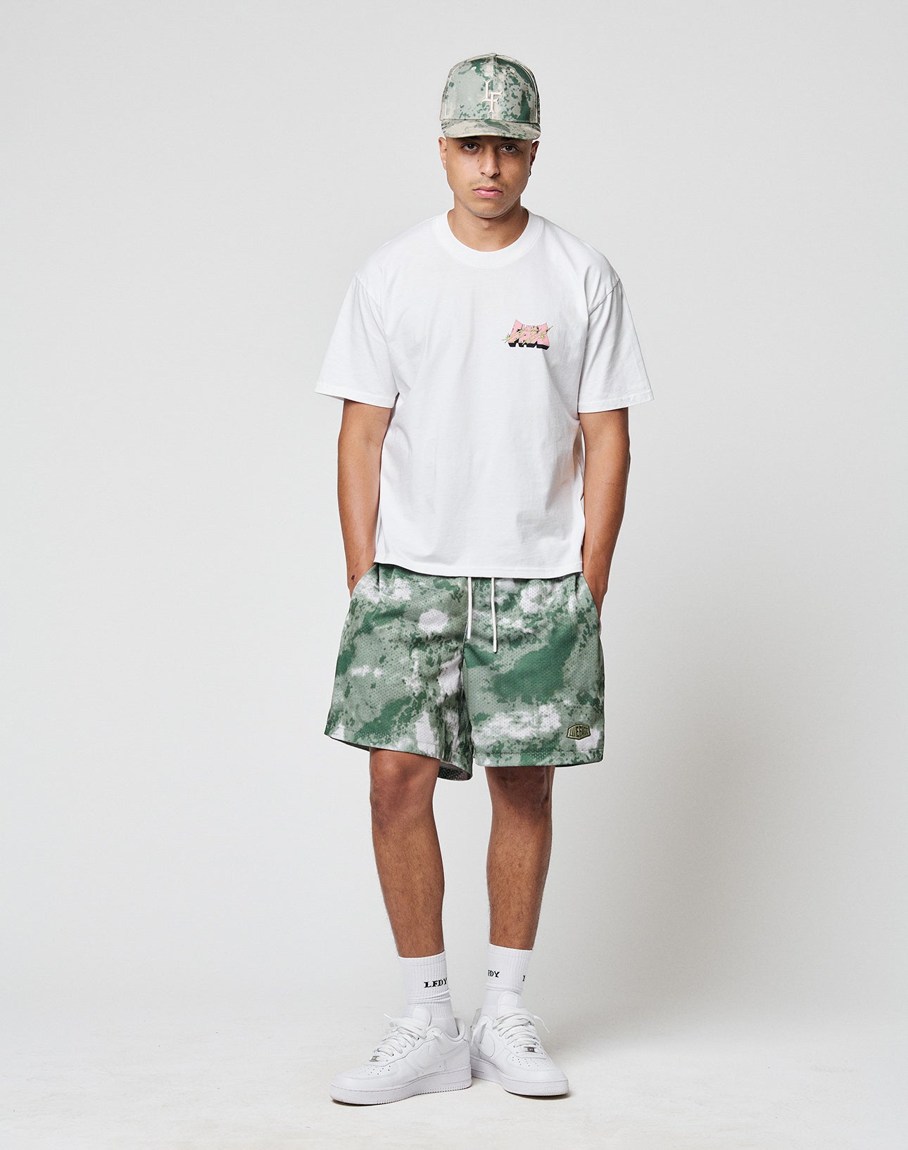 A man stands against a plain background wearing the LIVE FAST DIE YOUNG LFDY Thunder Chest Tee in white, paired with green and white tie-dye shorts, a matching cap, white crew socks, and sneakers. He looks straight ahead with hands in his pockets.
