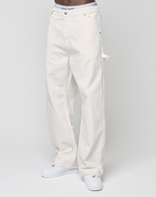 A person wearing LIVE FAST DIE YOUNG Worker Pants with a baggy fit and carpenter pocket, featuring a white waistband with "LIVE FAST" text, paired with white sneakers against a plain light background.