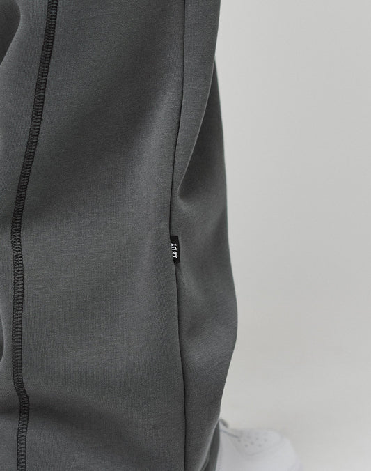 Close-up of a person wearing LIVE FAST DIE YOUNG LFDY Active Fleece Jogger in dark gray with black stitching and a small black fabric tag on the seam; part of a white sneaker is visible. The background is plain and light-colored.