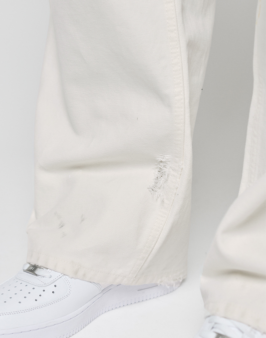 Close-up of a person wearing LIVE FAST DIE YOUNG Worker Pants in off-white with a baggy fit, carpenter pocket, and a small tear near the hem, paired with white sneakers against a plain light background.