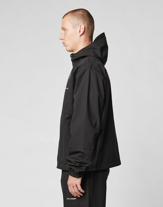 A man with short hair wears the LIVE FAST DIE YOUNG Basic Shell Jacket, featuring a water-repellent zipper, and black pants. He stands in profile against a plain light gray background.