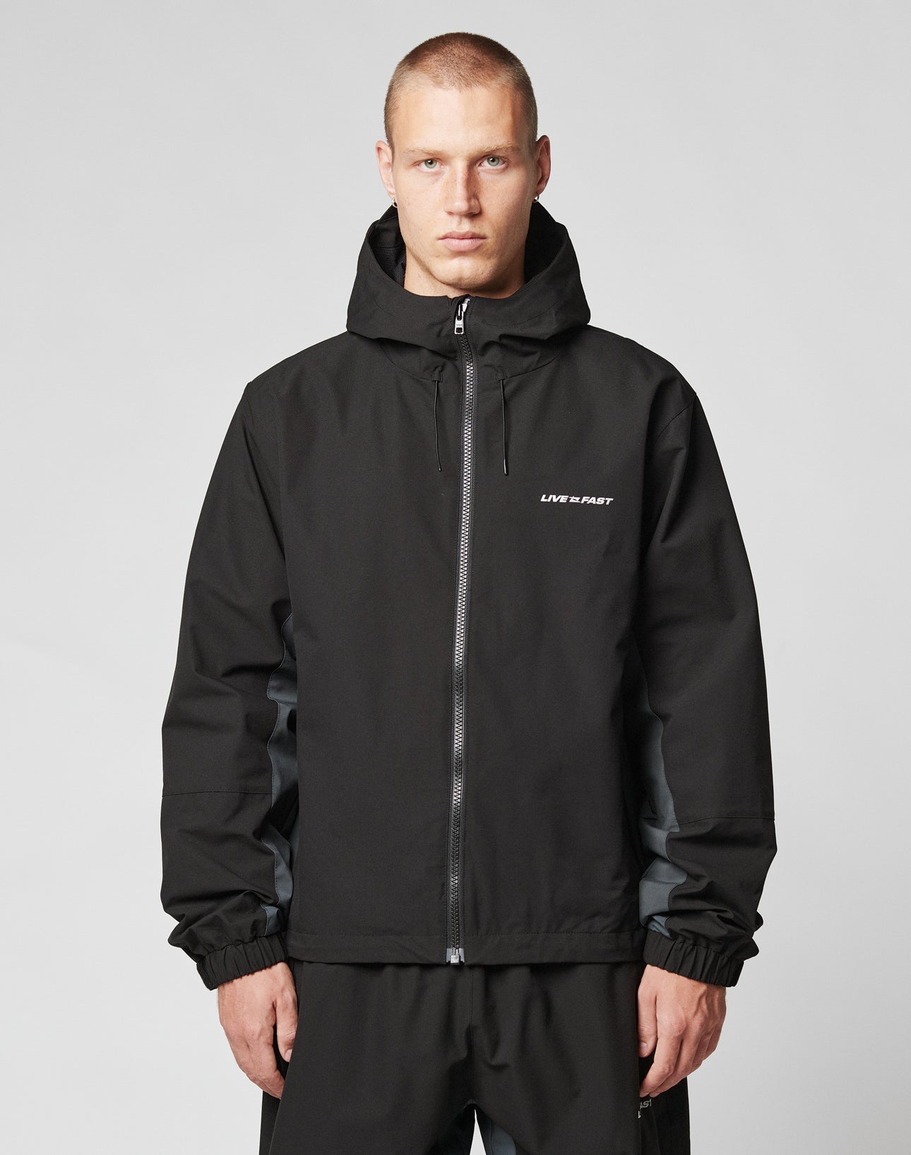 A short-haired person wears the LIVE FAST DIE YOUNG Basic Shell Jacket with reflektierender Druck and LIVE FAST on the chest, featuring an oversize fit, standing against a plain light gray background.