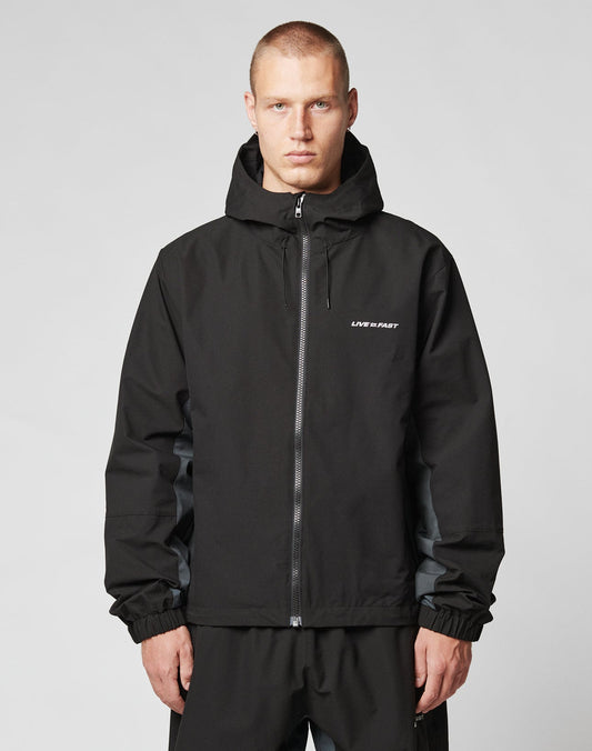 A short-haired person wears the LIVE FAST DIE YOUNG Basic Shell Jacket with reflektierender Druck and LIVE FAST on the chest, featuring an oversize fit, standing against a plain light gray background.