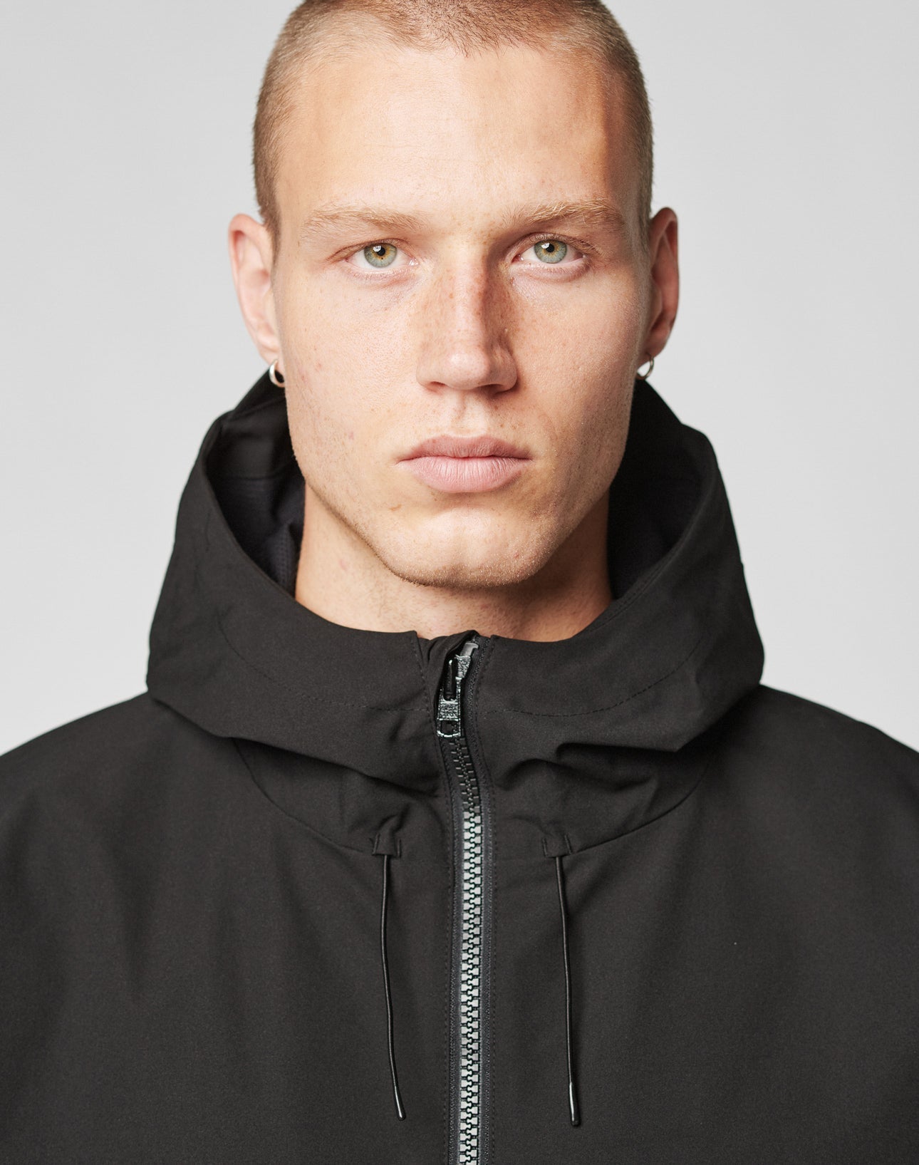 A person with short blonde hair and light eyes wears the LIVE FAST DIE YOUNG Basic Shell Jacket in black, oversize fit, zipped to the neck, staring at the camera against a plain light background.